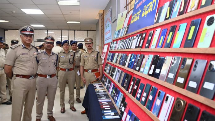 Police officers in uniform stand beside a wall displaying hundreds of mobile phones on red felt, likely a confiscated device exhibit.