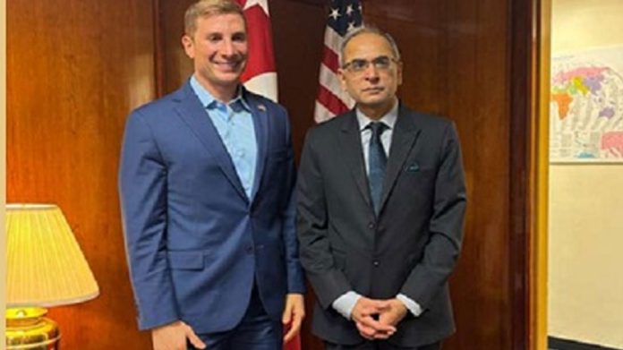 Two men in business suits pose for a photo in an office with an American flag in the background.