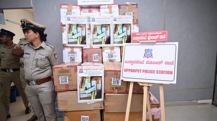 Uniformed police officers beside a tall stack of cardboard boxes and posters outside Upparpet Police Station.